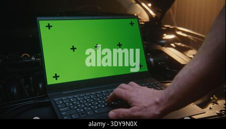 Chroma Key Laptop in Auto Workshop für Bildschirmaustausch. Laptop mit leuchtendem grünem Bildschirm und Tracking-Punkten über dem Motor, während die männliche Hand mit dem Tippen beginnt. Konzept der Inhaltserstellung. Stockfoto