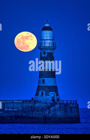 Ein Blick in der Abenddämmerung auf den March Worm Full Moon, der in einen klaren Himmel über dem Roker Pier und dem Leuchtturm in Sunderland, Tyne & Wear aufsteigt Stockfoto