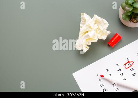Hochwinkelansicht einer zerknitterten Papierkugel, eines roten Markers und eines Kalenders mit eingekreistem Datum auf einer olivgrünen Oberfläche mit Kopierraum. Stockfoto
