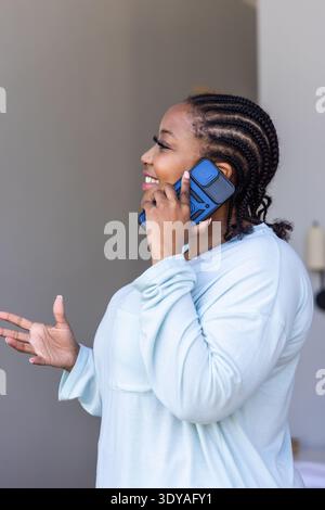 Afroamerikanerin lächelt und spricht auf blauem Smartphone, trägt hellblaues Oberteil zu Hause Stockfoto