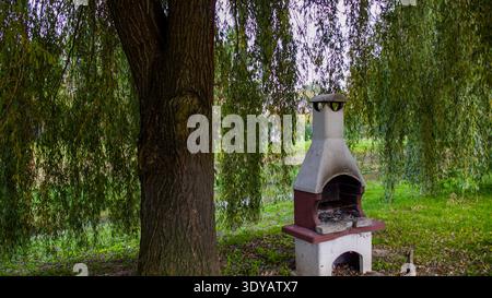Ein traditioneller Gartengrill aus Beton im Freien, der unter der großen Trauerweide in einem ländlichen polnischen Gebiet steht. Stockfoto
