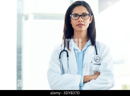 Überkreuzte Arme, Brille und Porträt des Arztes im Krankenhaus mit Vertrauen zur ärztlichen Betreuung. Ernsthafte, professionelle und weibliche Chirurgin mit Stolz auf Stockfoto