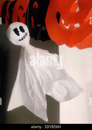 Hausgemachte Halloween-Geisterdekoration aus weißem Taschentuch mit gezeichneten schwarzen Augen und Mund, die an einer Schnur unter orangefarbenem und schwarzem Kürbis gar hängt Stockfoto