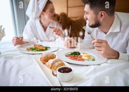 Glückliches Paar genießt Frühstück mit Hund auf dem Bett in einem gemütlichen Schlafzimmer Stockfoto