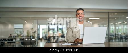Glücklicher Geschäftsmann mittleren Alters, der im Büro mit Laptop in die Kamera blickt. Kopierraum Stockfoto