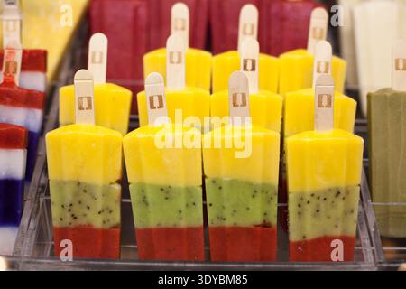Reihen von mehrschichtigen Fruchteis mit Mango-, Kiwi- und Erdbeeraromen. Holzstäbchen in gefrorene Leckereien. Anzeige in transparentem Kunststoff t Stockfoto