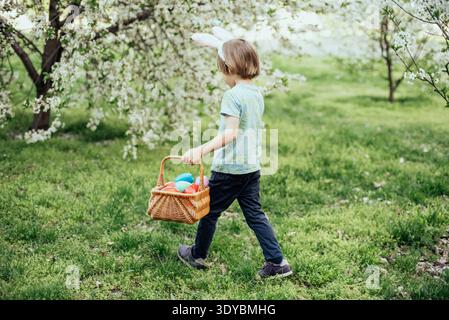 Ostereierjagd. Ein Kind mit Hasenohren läuft, um Ei im Garten abzuholen. Ostertradition. Baby mit Korb voller bunter Eier. Stockfoto