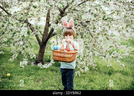 Ostereierjagd. Ein Kind mit Hasenohren läuft, um Ei im Garten abzuholen. Ostertradition. Baby mit Korb voller bunter Eier. Stockfoto