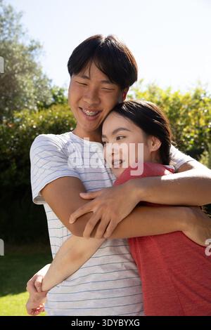 Asiatisches Paar umarmt auf dem Rasen im Garten unter hellem Sonnenlicht und zeigt Zahnspangen und Ohrringe. Romantik, Zuneigung, Outdoor, Lifestyle, Glück, lässig, Natur || Model veröffentlicht Stockfoto