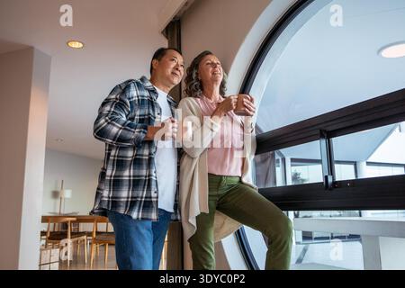 Verschiedene Paare, die Keramikbecher halten und durch ein kreisförmiges Fenster im Wohnbereich blicken. Modern, gemütlich, Lifestyle, Interieur, Ruhe, Ruhe, malerische Landschaft || Model veröffentlicht Stockfoto
