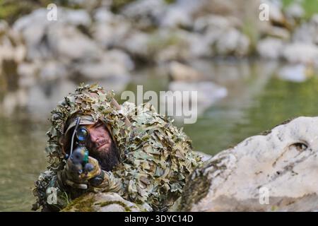 Scharfschütze mit Ghillie-Anzug und Tarnfarbe liegt auf einem felsigen Bach-Ufer und zielt mit einem Gewehr ab. Das Bild vermittelt Tarnung, Fokus, taktisches Training und militärische Aufklärung im Freien. || Modell freigegeben Stockfoto