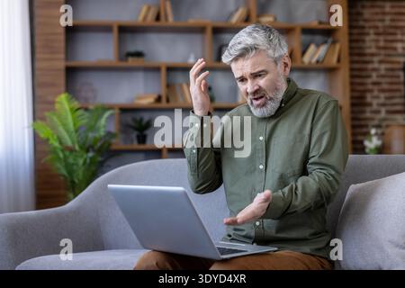 Reifer Mann mit grauen Haaren sitzt auf einem Sofa, frustriert und besorgt, während er zu Hause an einem Laptop arbeitet und mit Computerproblemen und Verbindungsproblemen zu kämpfen hat Stockfoto