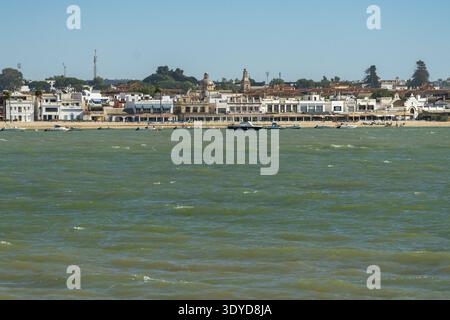Die Küstenstadt Sanlucar de Barrameda mit weißen Gebäuden, Kirchen und kleinen Booten auf dem grünlichen Meer unter klarem Himmel zeigt die Küstenstadt Sanlucar de Barrameda Stockfoto