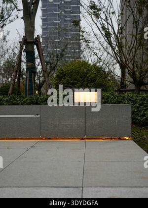 Glühende Außenlampe auf Steinsitzen mit Hochhäusern im Hintergrund bei Dämmerung. Das Bild vermittelt urbanen Abend und moderne Communi Stockfoto