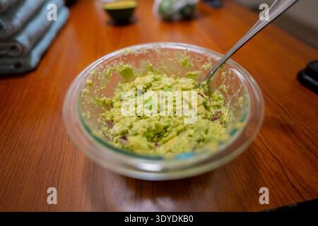 Frische hausgemachte Guacamole in klarer Glasschale auf Holztisch mit Löffel, gemahlene Avocadomischung für Tacos zubereitet, gesunde Kochszene in der Küche Stockfoto