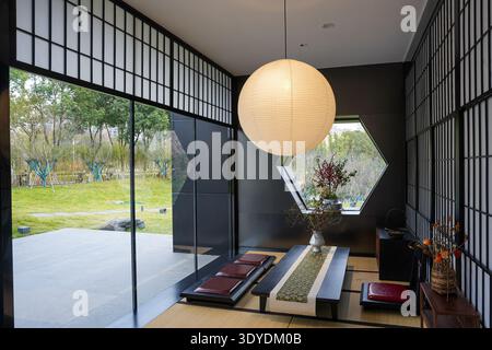 Zimmer im orientalischen Stil mit Tatami-Matten, niedrigem Holztisch, Bodenkissen und einer großen Pendelleuchte aus Papier durch ein sechseckiges Fenster. Die Bildkonve Stockfoto