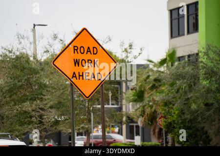Leuchtend orangefarbenes Schild FÜR STRASSENARBEITEN neben Bäumen und städtischen Gebäuden in den Vereinigten Staaten, das Straßeninstandhaltung, Verkehrswartung und Straßenverkehr hervorhebt Stockfoto