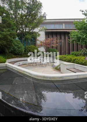 Ein eleganter Außenbereich mit weißen Möbeln, Steinpflaster und einem ruhigen reflektierenden Wasserbecken in einem modernen Wohngarten. Das Bild vermittelt sophist Stockfoto