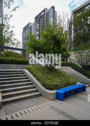 Eine elegante, moderne Steintreppe schlängelt sich durch einen lebendigen Garten in einem Wohngebiet, der von einer hellblauen Bank akzentuiert wird. Das Bild vermittelt eine strukturelle Darstellung Stockfoto