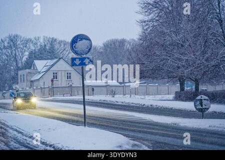 Ein Auto mit Scheinwerfern fährt im Winter auf einer schneebedeckten Straße, während der Schnee stark fällt und eine kalte Atmosphäre erzeugt. Stockfoto