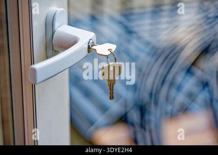 Metallschlüssel, die an einem weißen Türgriff hängen, mit einem unscharfen Hintergrund mit blauen und weißen Mustern, die Sicherheit und Zugang symbolisieren. Stockfoto