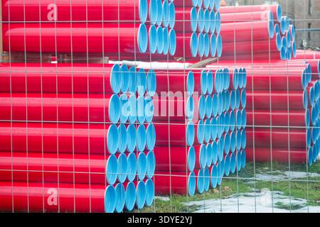Rote Kunststoffrohre mit blauen Enden sind in organisierten Reihen hinter einem Metallzaun auf einem grasbewachsenen Außenbereich mit Schneeflächen gestapelt. Stockfoto