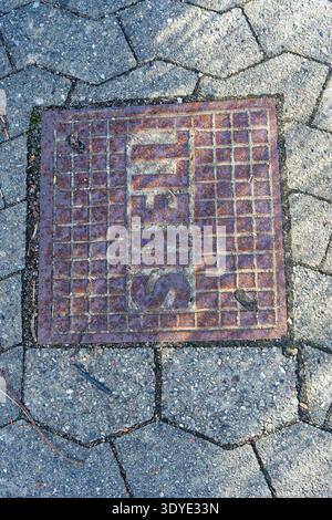 Eine verwitterte, rostige Metallkanalabdeckung, eingebettet in einen strukturierten Steinpflaster mit sechseckigen Fliesen und kleinen Trümmern, die herum verstreut sind. Stockfoto