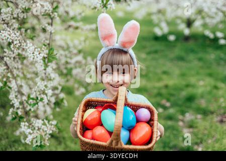 Ostereierjagd. Ein Kind mit Hasenohren läuft, um Ei im Garten abzuholen. Ostertradition. Baby mit Korb voller bunter Eier. Stockfoto