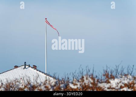 Ein schneebedecktes Dach mit Schornsteinen und eine hohe dänische Flagge Danebrog Dannebrog mit einem roten Windsock vor einem klaren blauen Himmel an einem kalten Wintertag. Stockfoto