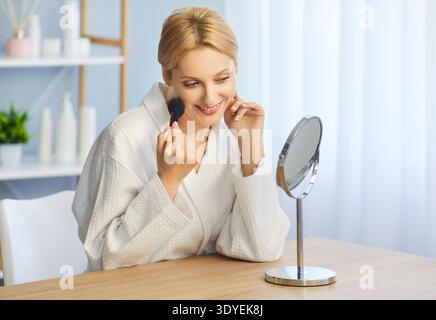 Blonde Frau, die Make-up mit dem Pinsel vor dem Spiegel aufträgt Stockfoto