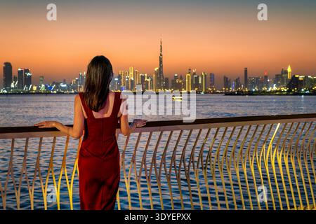 Rückansicht einer wunderschönen Frau in einem roten Abendkleid, die in der Abenddämmerung die ferne Skyline von Dubai, VAE, betrachtet Stockfoto