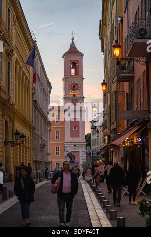 Uhrenturm in der Nähe des Palais Rusca, Nizza, Frankreich. Fußgänger gehen an bunten Gebäuden und beleuchteten Laternen in der historischen Altstadt vorbei. Stockfoto