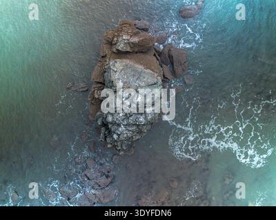 Drohnenansicht einer zerklüfteten Felsformation, die aus dem ruhigen tiefblauen Meereswasser hervorgeht. Wellen erzeugen weiße Schaummuster rund um die isolierte Insel Stockfoto