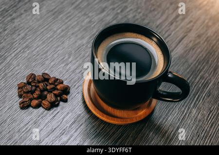 Schwarze Tasse Kaffee gefüllt mit dunklem Kaffee mit Kaffeebohnen, alles auf einem grauen Tisch - gemütliche Frühstücksszene Stockfoto