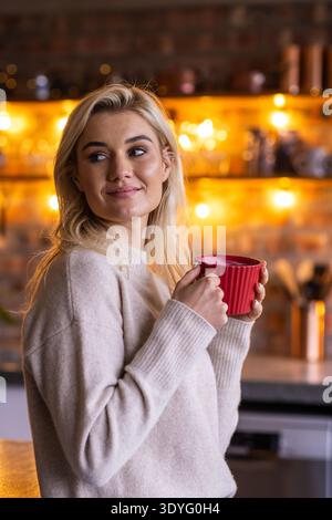 Erwachsene Frau mit cremefarbenem Pullover, der rote Becher mit beiden Händen hält und links in der Küche schaut Stockfoto