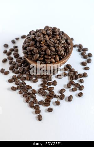 Ein vertikales Hochwinkel-Foto von hochwertigen dunkel gerösteten Kaffeebohnen. Eine kleine rustikale Holzschale wird mit Bohnen gefüllt und auf einen sauberen weißen Sur gelegt Stockfoto