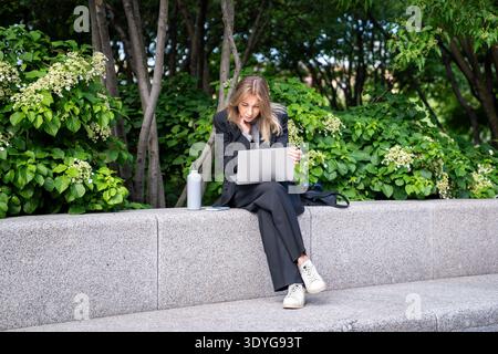 Durchdachte positive Geschäftsfrau, die im Stadtpark an einem Laptop arbeitet, Vorteile moderner Fernarbeit. Stockfoto