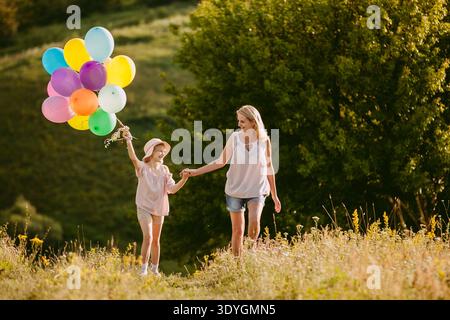 Frau mittleren Alters, die Hände hält mit einem fröhlichen Mädchen, das bunte Ballons trägt, auf einer sonnigen Wiese mit üppigem Grün spaziert. Stockfoto
