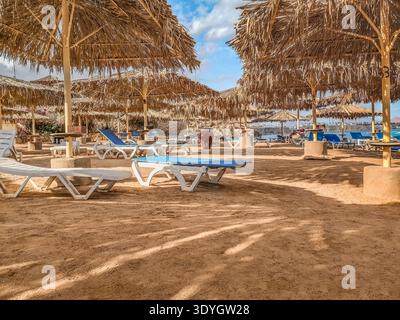 Strand am Roten Meer in Scharm El Sheikh, Ägypten. Leere Sonnenliegen unter Strohschirmen in einem tropischen Resort Stockfoto