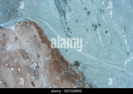 Komplizierte Texturen eines gefrorenen Feldes, wo trockenes braunes Gras auf mattierte blaue Erde trifft, die durch einen natürlichen Pfad geteilt wird. Dieser minimalistische Winterhintergrund fängt den ruhigen Übergang der Saison ein. Stockfoto