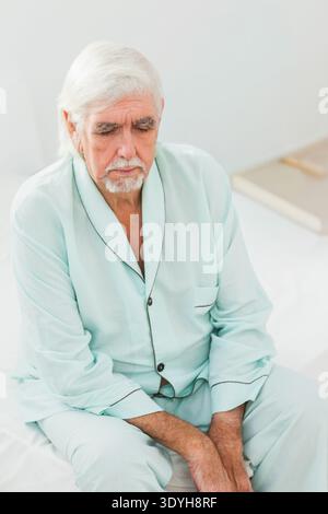 Ein älterer Mann sitzt auf dem Bett und trägt einen blauen Pyjama im Schlafzimmer. Er blickt nach unten, Hände auf Knien. Nachtwäsche, minimalistisch, besinnlich, ruhig, weich, d Stockfoto