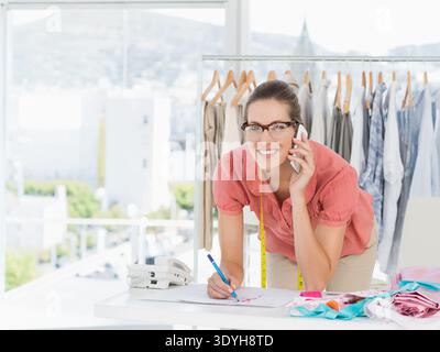 Frau aus den 30er Jahren, die Bluse und Maßband trug, auf Papier skizzierte und im Studio mit Smartphone sprach. Designer, Atelier, Stoffe, Farbmuster, Muster, dra Stockfoto