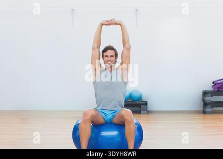 Mann sitzt auf blauem Trainingsball, dehnt sich über dem Studio aus, trägt sportliche Oberteile und Shorts. Stabilität, Fitnessstudio, Fitness, Balance, lila, Matten, ste Stockfoto
