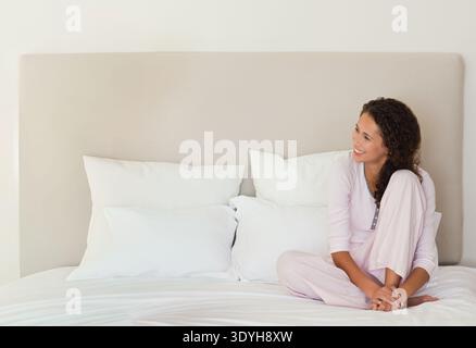 Erwachsene Frau, die im Schlafzimmer mit Kissen und Kopfteil auf einem Kingsize-Bett sitzt und Pyjama trägt, Kopierraum. Nachtwäsche, ruhig, entspannt, ruhig, gemütlich, neutral, Meilen Stockfoto