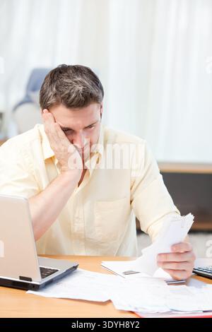 Mann im mittleren Erwachsenenalter, der am Holzschreibtisch im Heimbüro sitzt und Dokumente in der Nähe eines offenen silbernen Laptops hält. Kontemplation, Konzentration, introspektiv, Fokus, wo Stockfoto