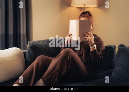 Eine junge Frau sitzt zu Hause bequem auf einem blauen Sofa und hält ein Buch vor ihrem Gesicht, während sie unter warmem Lampenlicht liest, um sich zu entspannen Stockfoto