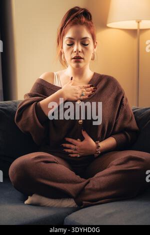 Eine junge rothaarige Frau sitzt im Kreuz auf einem blauen Sofa in einem gemütlichen Wohnzimmer und übt somatische Atemmeditation mit den Händen auf ihrem Herzen A Stockfoto