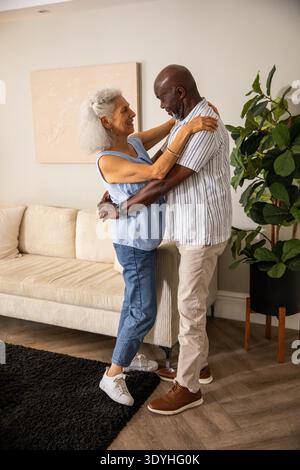 Vielseitiges Seniorenpaar, das sanft im Wohnzimmer mit abstrakter Wandmalerei und Topfpflanze tanzt. Romantik, Gesellschaft, Gelassenheit, Eleganz, Innenraum, Lifestyle, Wertschätzung || Model veröffentlicht Stockfoto