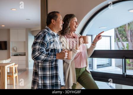 Verschiedene Paare stehen im Wohnbereich mit einem bogenförmigen Fenster, das Keramikbecher hält und nach draußen blickt. Modern, warm, ästhetisch, Freizeit, Verbindung, Ruhe, gemütlich || Model veröffentlicht Stockfoto