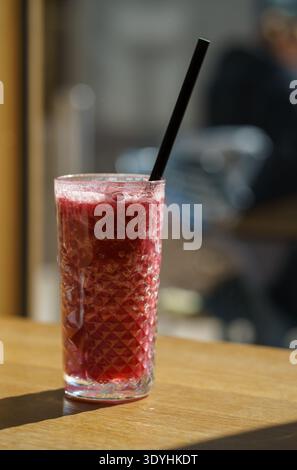 Erfrischender Smoothie mit roten Beeren, Eis in strukturiertem Glas und Strohhalm auf Holztisch. Gesundes Fruchtgetränk wird drinnen in einem gemütlichen Café mit sanftem Licht serviert Stockfoto